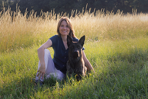 Person sitzt im Gras. Neben ihr ein liegender Hund.